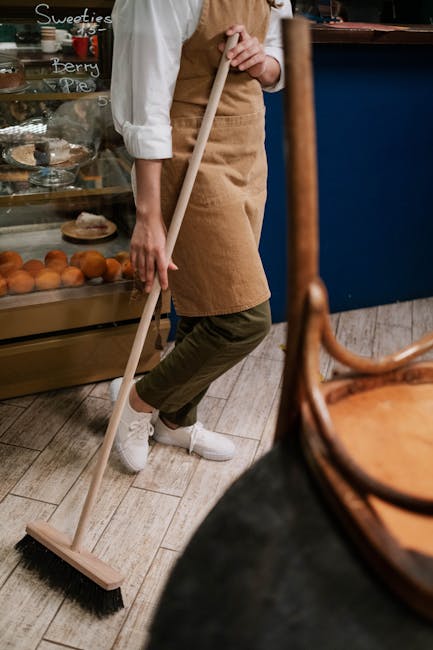 Close-up of a person sweeping a wooden or laminate floor in a commercial or domestic setting, with a focus on their legs in beige apron and white sneakers, using a push broom. The setting includes a glass display case with baked goods and a chalkboard menu in the background, indicating a café or bakery environment. The floor appears clean and well-maintained, reflecting an emphasis on surface cleaning and hygiene, as part of regular sanitation and domestic cleaning practices. Islington Carpet Cleaners offers professional cleaning services to maintain such cleanliness, ensuring surfaces are dust-free and sanitized. Proper sweeping contributes to a hygienic space, aligned with advice on avoiding hidden cleaning charges by keeping surfaces well-maintained.