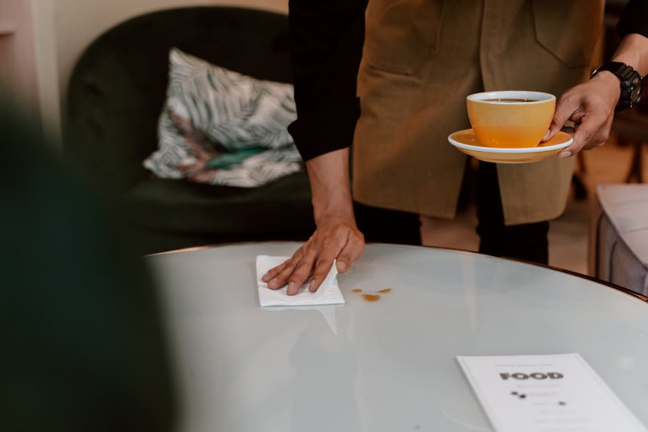 A person wearing a black long-sleeve shirt and brown apron is wiping a smooth, glossy white countertop with a white cloth, removing coffee stains. They are holding a white saucer with an orange-rimmed ceramic cup filled with coffee in their other hand. The background features a dark-colored sofa with a decorative pillow that has a leaf pattern, and soft, warm lighting enhances the clean and tidy appearance of the domestic setting. The image emphasizes surface cleaning and maintenance, aligning with professional cleaning practices that Islington Carpet Cleaners offers for residential spaces, focusing on hygiene and spotless surfaces.