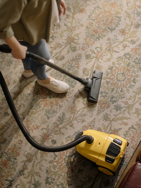 A child using a vacuum cleaner to thoroughly clean an ornate, patterned carpet in a domestic living space. The carpet features a floral design in muted tones of beige, green, and rust, indicative of Victorian style often found in Islington homes. The vacuum cleaner, positioned centrally, has a yellow body with a black hose and attachment, while the child is dressed in casual clothing with beige sneakers and blue jeans. Soft ambient lighting illuminates the room, highlighting the cleanliness and well-maintained condition of the surface. This scene exemplifies routine domestic cleaning and surface hygiene, which is part of general carpet maintenance carried out by professional cleaning company Islington Carpet Cleaners, especially relevant for addressing access issues in period homes with intricate flooring.