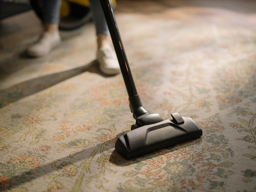 A child using a vacuum cleaner to thoroughly clean an ornate, patterned carpet in a domestic living space. The carpet features a floral design in muted tones of beige, green, and rust, indicative of Victorian style often found in Islington homes. The vacuum cleaner, positioned centrally, has a yellow body with a black hose and attachment, while the child is dressed in casual clothing with beige sneakers and blue jeans. Soft ambient lighting illuminates the room, highlighting the cleanliness and well-maintained condition of the surface. This scene exemplifies routine domestic cleaning and surface hygiene, which is part of general carpet maintenance carried out by professional cleaning company Islington Carpet Cleaners, especially relevant for addressing access issues in period homes with intricate flooring.