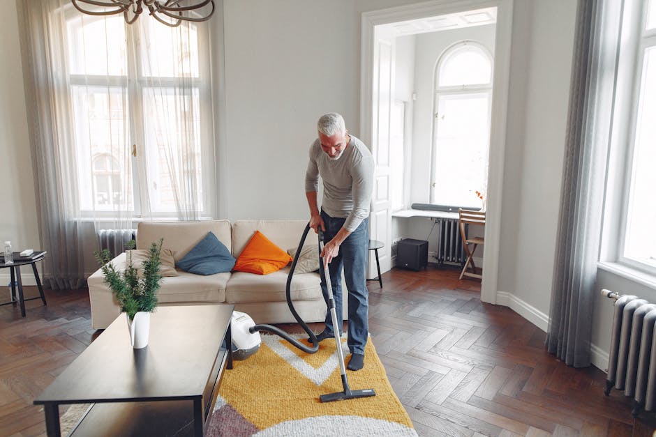 A person kneeling on a patterned carpet in a domestic setting, preparing a yellow portable vacuum cleaner for surface cleaning. The individual is wearing a beige blouse and blue jeans, and is opening the vacuum's dust compartment or preparing it for use. The carpet has a traditional floral design with earthy tones, and the room features warm lighting, highlighting the cleanliness of the carpet surface. The scene illustrates a routine deep cleaning process associated with domestic cleaning services offered by Islington Carpet Cleaners, emphasizing thorough removal of dust and debris from home flooring surfaces.