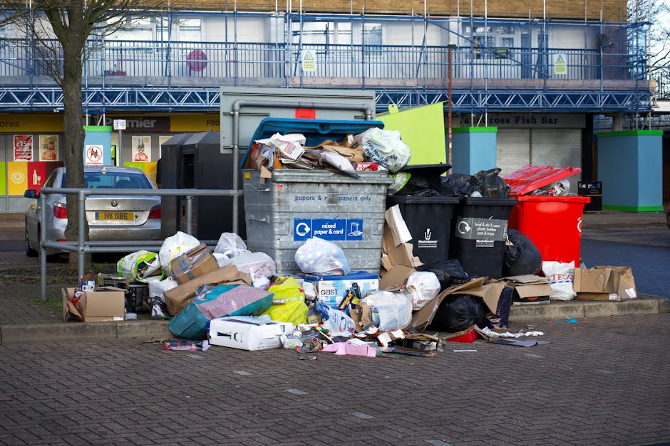 A roadside collection of overflowing waste bins and discarded rubbish including cardboard boxes, paper bags, plastic packaging, and black trash bags, situated on a paved sidewalk in front of a commerc