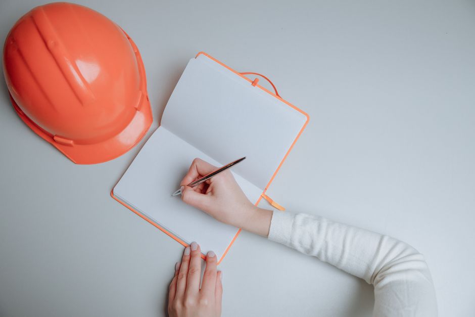 A person's hand holding a silver pen is positioned above an open, blank notebook with an orange border, placed on a light grey surface. To the left of the notebook, there is an orange safety helmet re