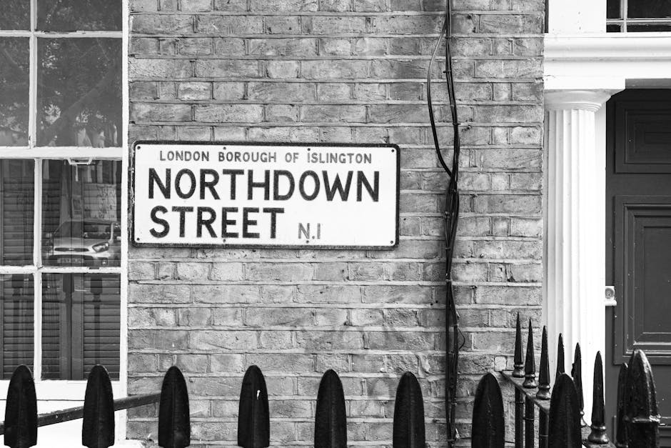 A black-and-white photograph of a street sign reading 'Northdown Street' mounted on a brick wall in Islington, London, with the top of a window visible to the left and a set of black iron railings at the bottom. The brick wall appears clean and well-maintained, with visible mortar lines, and the sign is centered in the image. The area is illuminated by natural light, highlighting the textures of the brick and metal, and there's an electrical cable running vertically beside the sign. This image captures the urban street scene typical of Islington, emphasizing the classic brickwork and street signage.