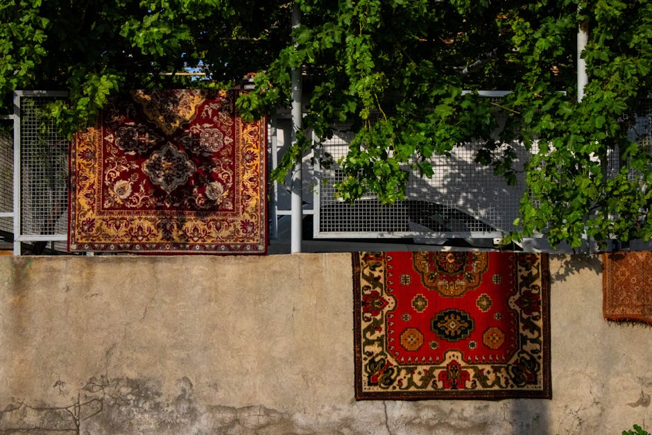 An outdoor balcony area featuring two ornate, patterned rugs with intricate floral and geometric designs in red, black, and beige tones, placed on a concrete floor with signs of wear and some discoloration. The balcony has a metal railing and is partially shaded by green foliage from a climbing plant or vine, creating a dappled shadow across the rugs. The sunlight highlights the vibrant colors of the rugs and the lush greenery surrounding the space. The scene suggests a well-maintained, decorative outdoor area where Islington Carpet Cleaners might perform surface cleaning or deep cleaning services to preserve the rugs' appearance and hygiene, especially in an environment exposed to outdoor elements.