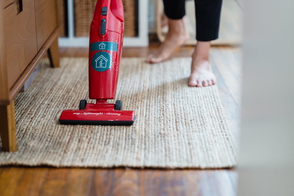 A red handheld vacuum cleaner resting on a beige carpet in a residential room with hardwood flooring, partially visible wooden furniture, and a person barefooted standing on the carpet. The vacuum appears to be in use or ready for surface cleaning, with a focus on maintaining hygiene and tidiness in the home. The room is well-lit, highlighting the clean and orderly surface, aligning with the services offered by Islington Carpet Cleaners on their page about same day carpet cleaning delays and common problems in Islington.