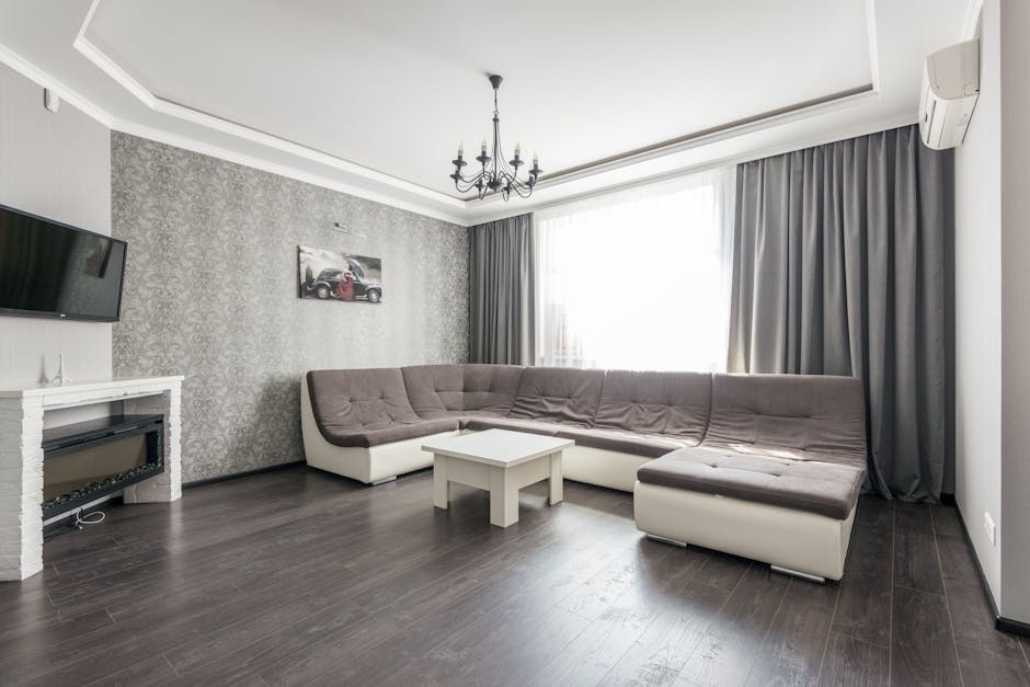 A modern living room with a large L-shaped sectional sofa covered in light brown fabric, positioned against a textured silver-grey wallpapered wall. A small white coffee table is placed in front of the sofa. To the left, a flat-screen TV is mounted on the wall above a white fireplace mantel, and an electric fireplace is installed beneath it. The room features dark wooden flooring, brightened by natural light coming through a large window dressed with grey curtains. A black chandelier hangs from the white ceiling, and an air conditioning unit is mounted on the right wall. The surfaces appear clean and well-maintained, suitable for routine domestic surface cleaning and sanitisation, as provided by Islington Carpet Cleaners.