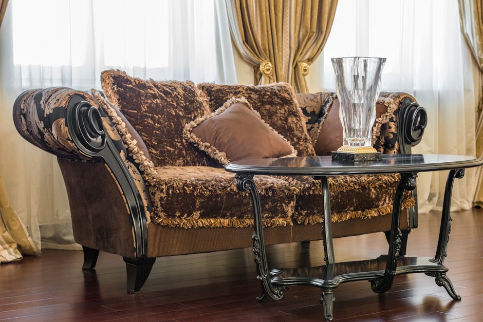 A plush, vintage-style sofa with ornate black wooden arms and floral upholstery fabric, situated in a well-lit living room with sheer white curtains and gold drapes. The sofa is decorated with several cushions in shades of brown and beige, showing a soft and clean appearance. In front of the sofa is a dark wood coffee table with curved legs, holding a tall, faceted glass vase. The room features polished hardwood flooring, and the overall setting appears tidy and well-maintained, reflecting surface cleaning and domestic hygiene practices. The setting showcases a classic interior layout, emphasizing the importance of furniture sanitisation and deep cleaning by Islington Carpet Cleaners as part of their upholstery cleaning services for Canonbury and Barnsbury flats.