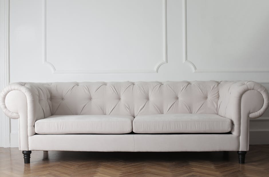 A modern living room with a large L-shaped sectional sofa covered in light brown fabric, positioned against a textured silver-grey wallpapered wall. A small white coffee table is placed in front of the sofa. To the left, a flat-screen TV is mounted on the wall above a white fireplace mantel, and an electric fireplace is installed beneath it. The room features dark wooden flooring, brightened by natural light coming through a large window dressed with grey curtains. A black chandelier hangs from the white ceiling, and an air conditioning unit is mounted on the right wall. The surfaces appear clean and well-maintained, suitable for routine domestic surface cleaning and sanitisation, as provided by Islington Carpet Cleaners.